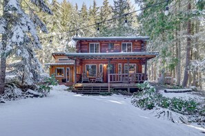 A log cabin with a porch and a deck, surrounded by snow-covered trees. - Rustic Sequim Cabin w/ Fire Pit & Forested Views! (Sequim)