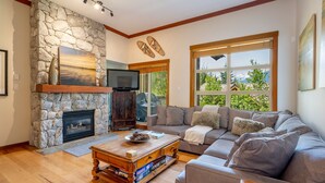A cozy living room with a stone fireplace, a large window with a view, and a comfortable gray sofa. - Ski-In/Ski-Out | Mountain Views | Private Hot Tub | Modern 3BR | Managed by Aloha Whistler (Whistler)