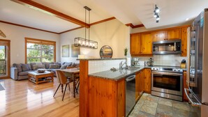 A spacious living area with a wooden floor, a large sofa, and a dining table. The kitchen features wooden cabinets, a stainless steel refrigerator, and a built-in oven. - Ski-In/Ski-Out | Mountain Views | Private Hot Tub | Modern 3BR | Managed by Aloha Whistler (Whistler)