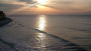 Une plage avec un coucher de soleil sur l’océan, un bâtiment sur la gauche et une mer calme.