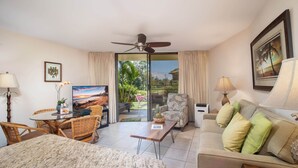 Habitación de hotel con cama, una mesa de comedor con sillas, un sofá, televisor y un balcón con vistas a palmeras y al atardecer.