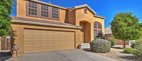 A two-story house with a beige exterior, a garage, and a well-maintained front yard with a small tree and shrubbery.