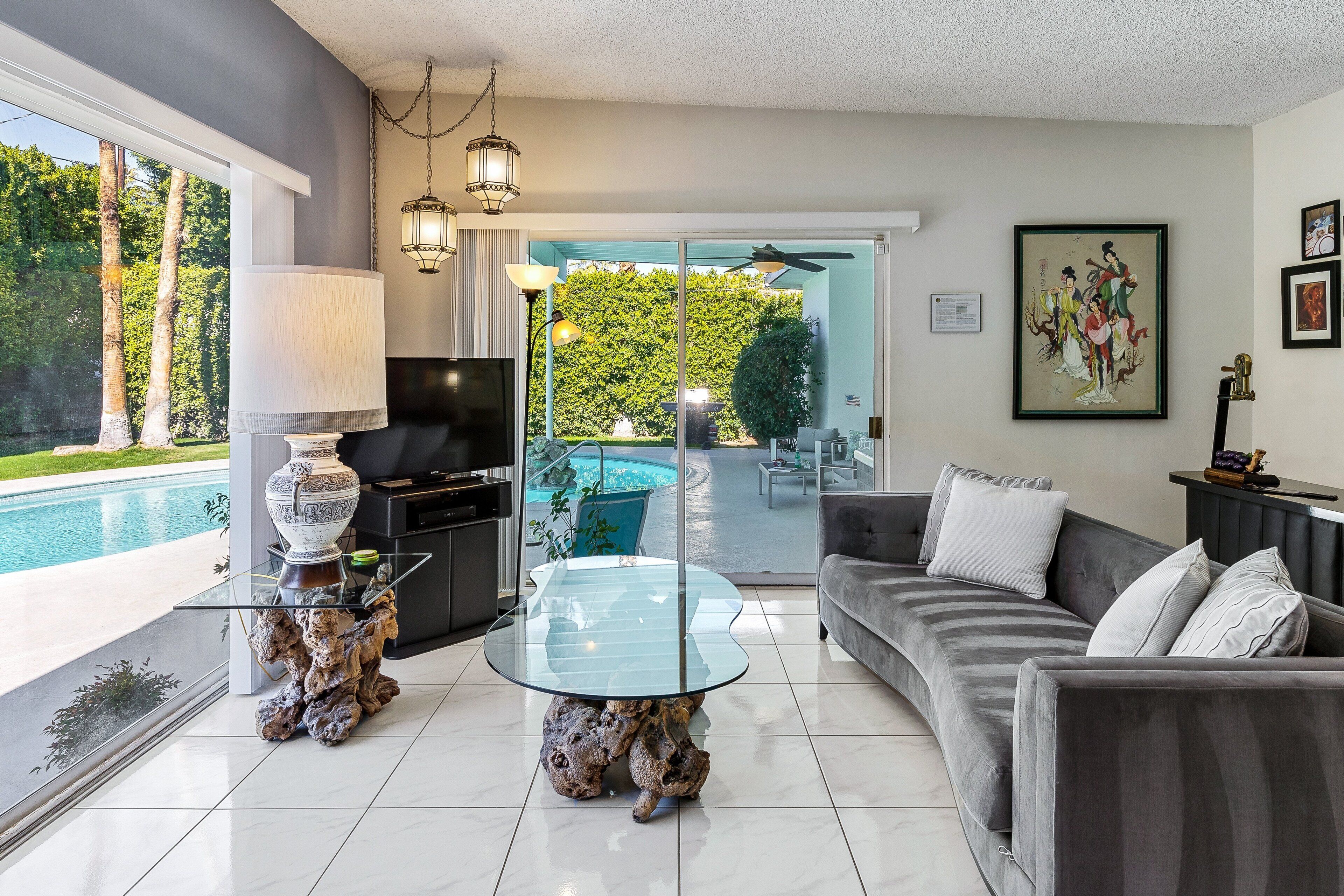 A modern living room with a glass coffee table, a sofa, a TV, and a view of a swimming pool.