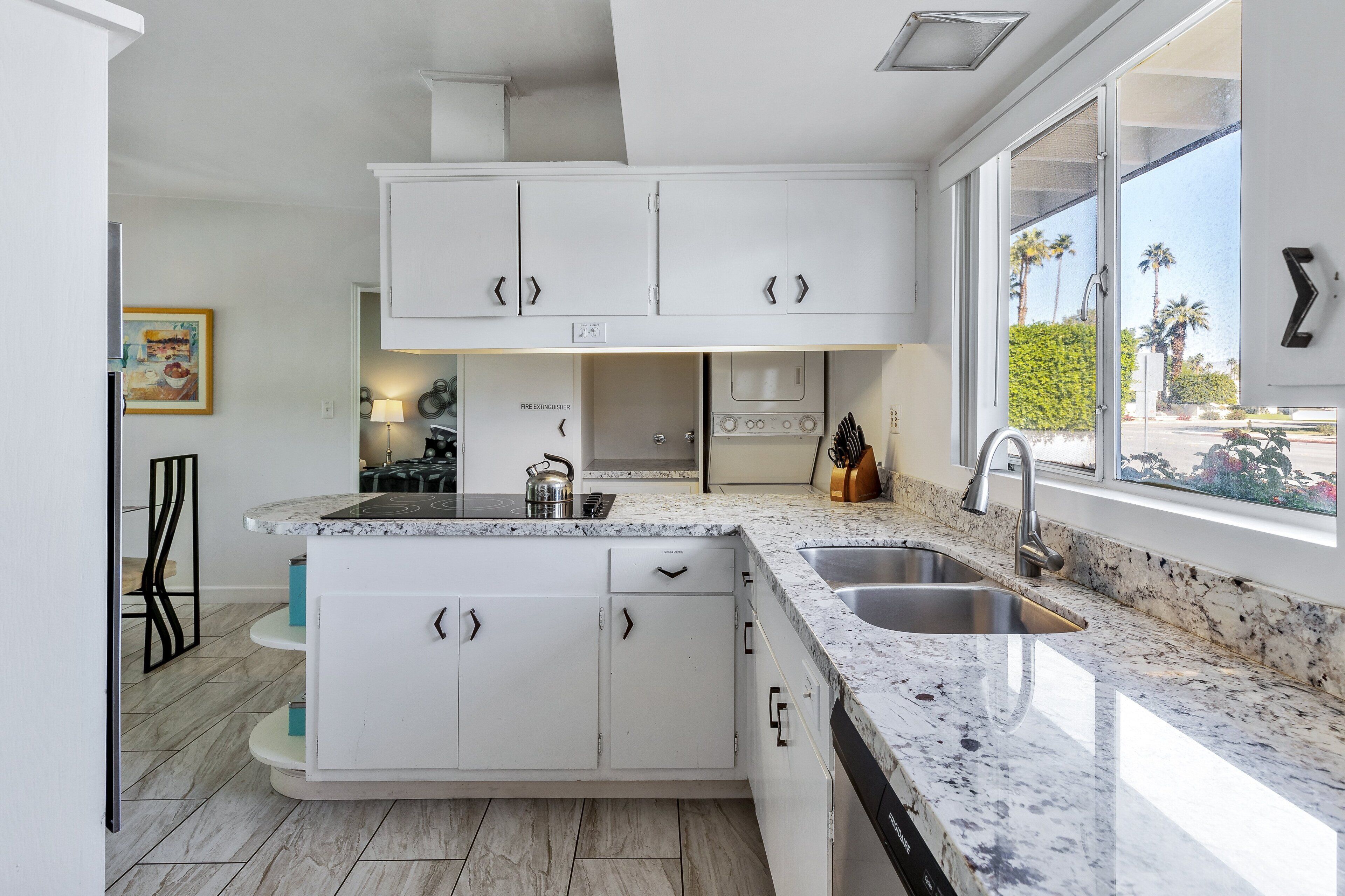A modern kitchen with white cabinetry, a granite countertop, a built-in stove, and a large window with a view of palm trees.