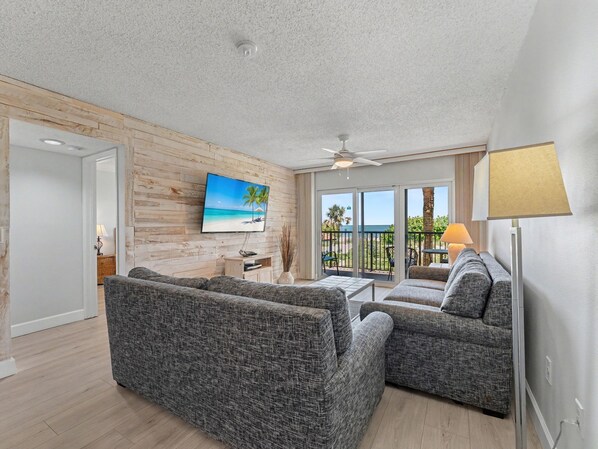 A living room with a TV, two sofas, a ceiling fan, and a view of the beach through large windows.