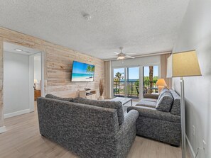 A living room with a TV, two sofas, a ceiling fan, and a view of the beach through large windows.