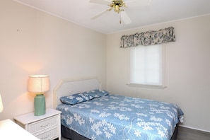 Une chambre à coucher avec un lit, une table de chevet avec une lampe, un ventilateur de plafond et une fenêtre avec des stores.