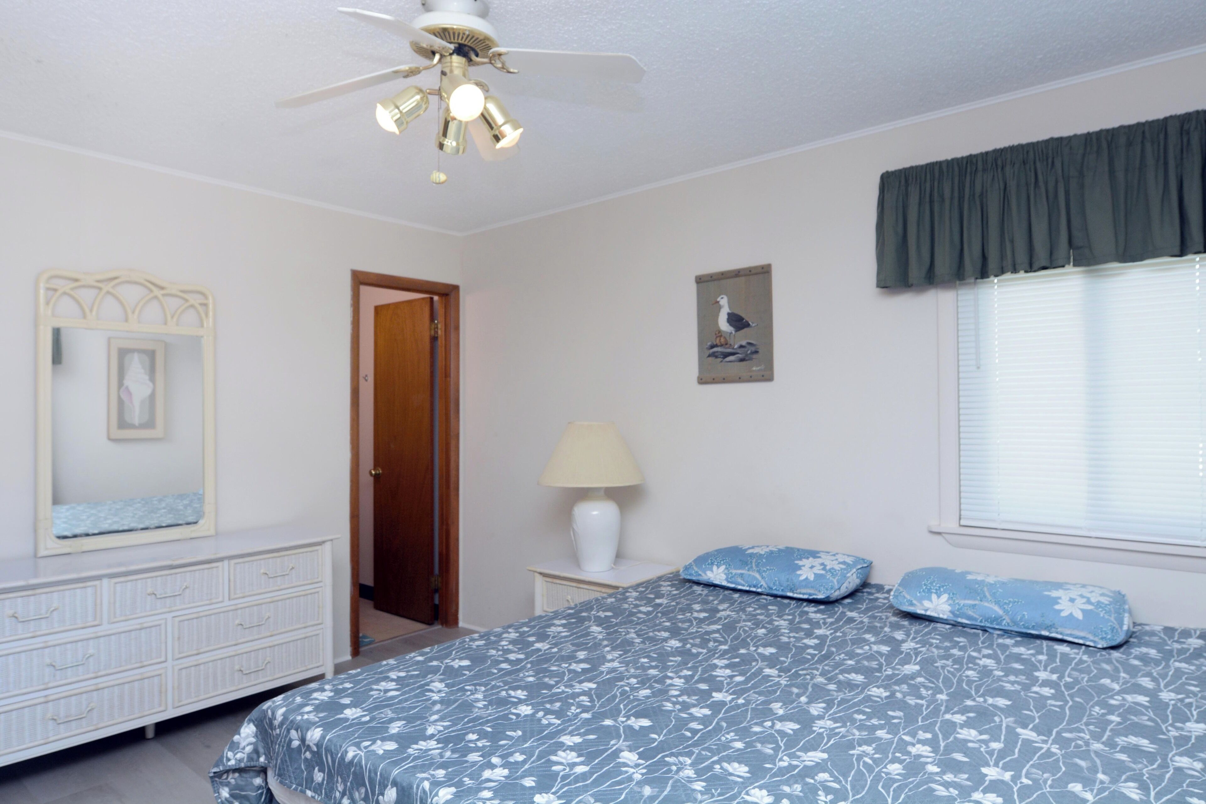 Ein Schlafzimmer mit Bett, Schminktisch, Lampe, Deckenventilator, einer Tür, einem Fenster mit Jalousien und einem Wandbild.