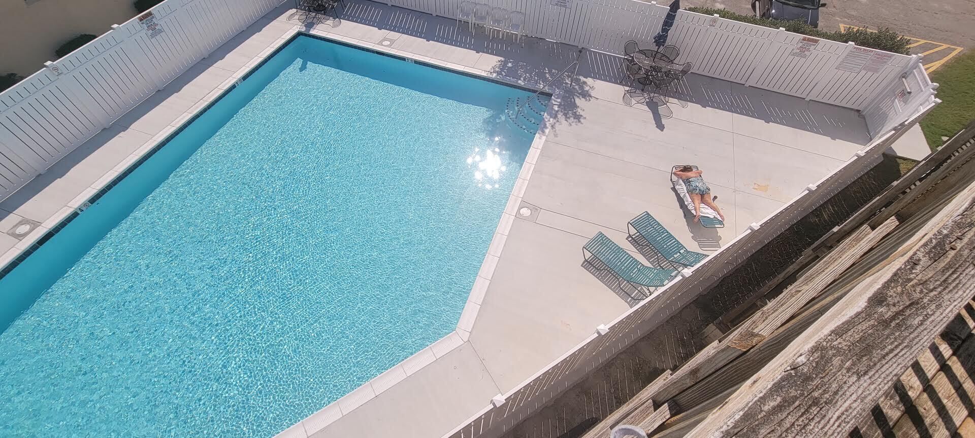 Uma piscina retangular com água azul cristalina, rodeada por um deck branco e uma cerca de madeira. Há espreguiçadeiras e uma mesa no deck.