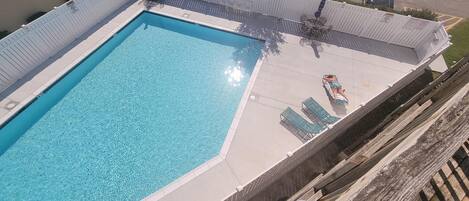 Uma piscina retangular com água azul cristalina, rodeada por um deck branco e uma cerca de madeira. Há espreguiçadeiras e uma mesa no deck.