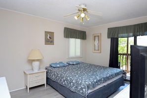 Une chambre à coucher comprenant un lit, une table de chevet, une lampe, un ventilateur de plafond et une fenêtre avec des rideaux.