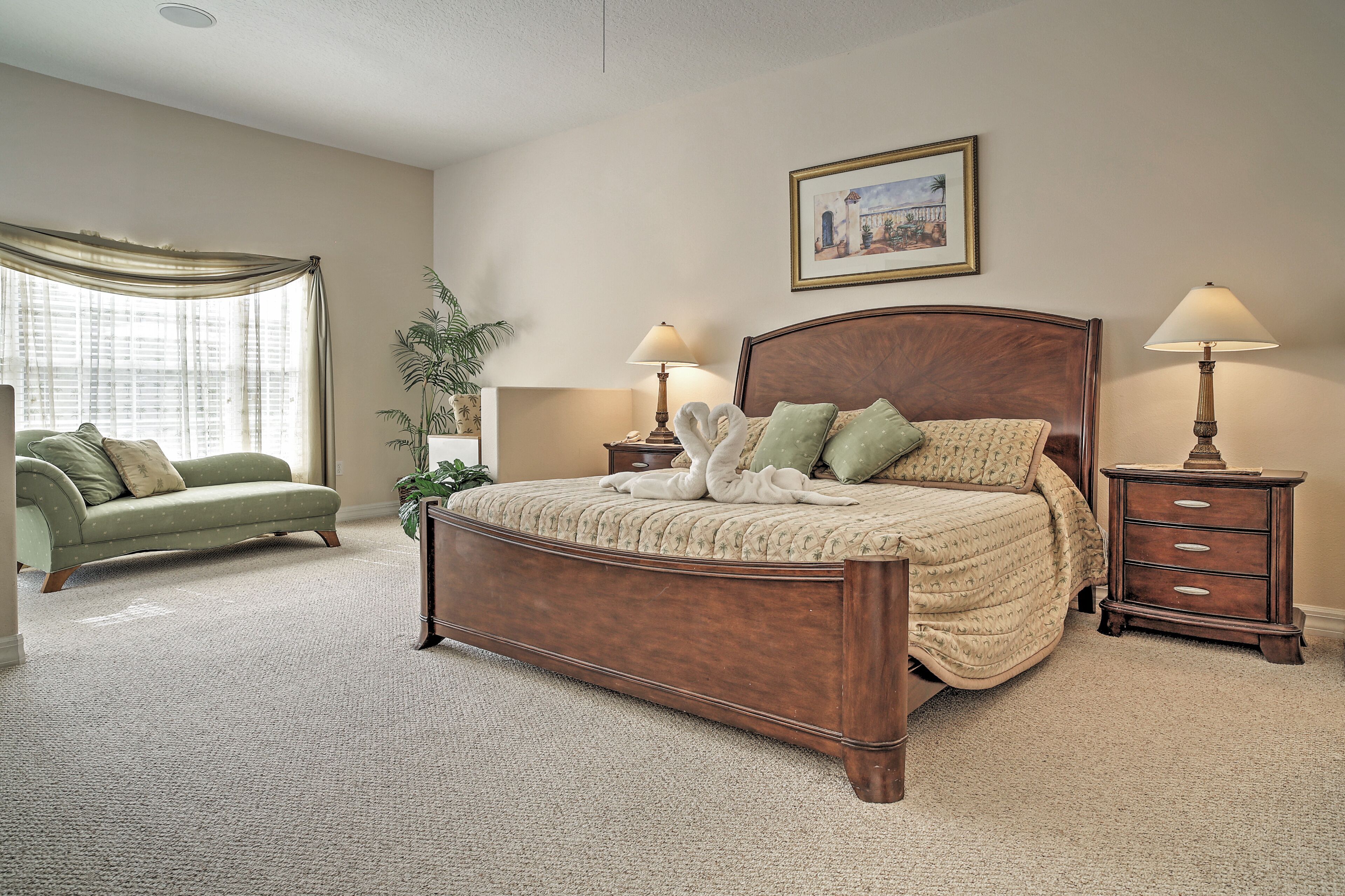 A bedroom with a large bed, two bedside tables with lamps, a green armchair, a window with curtains, and a framed picture on the wall.