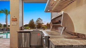 Una cocina exterior con una parrilla integrada, un refrigerador de acero inoxidable y un cartel que indica que el bar está abierto.