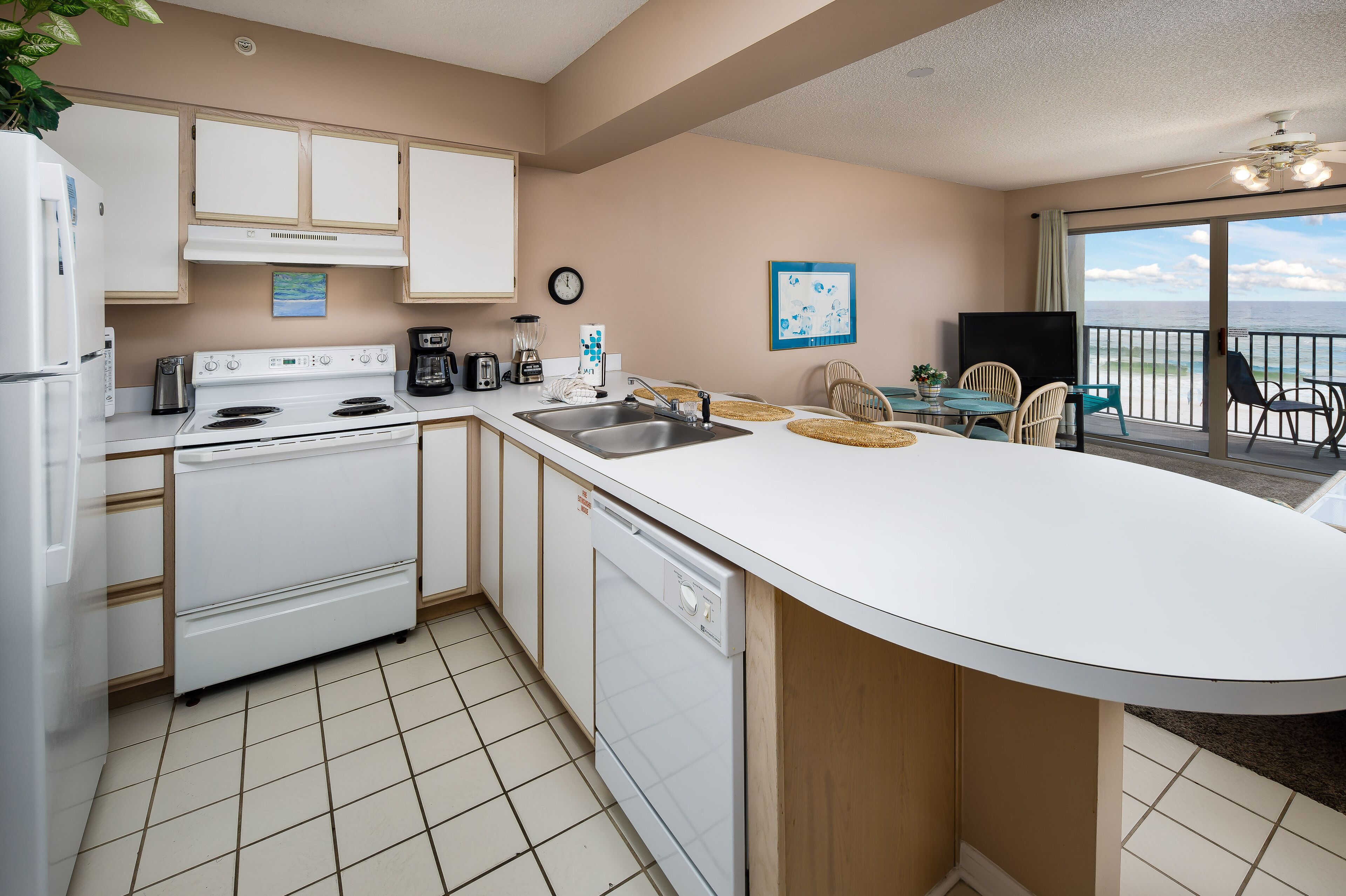 A kitchen with a white stove, dishwasher, and sink. There is a clock on the wall and a window with a view of the ocean.