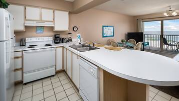 A kitchen with a white stove, dishwasher, and sink. There is a clock on the wall and a window with a view of the ocean.