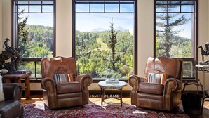 Une pièce spacieuse avec de grandes fenêtres donnant sur les montagnes, des fauteuils en cuir, une petite table ronde et un tapis à motifs.