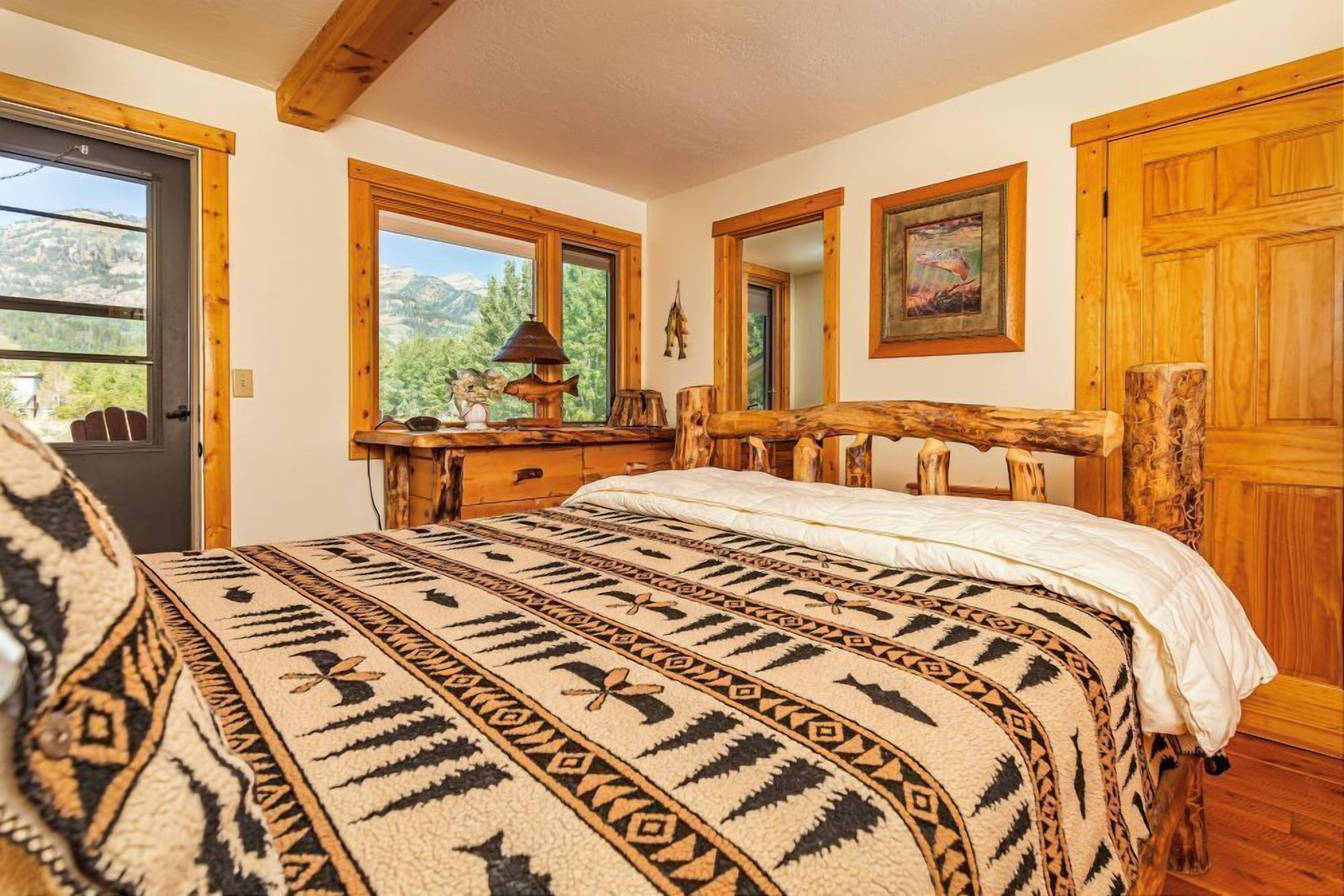 A bedroom with a bed, wooden headboard, a window with a view of mountains, a wooden dresser, and a lamp.
