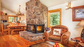 A cozy living room with a stone fireplace, wooden furniture, and a large window overlooking greenery.