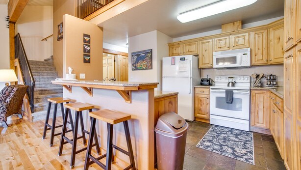 A kitchen with wooden cabinets, a white refrigerator, a microwave, and a stove. There is a wooden bar counter with stools and a trash can.