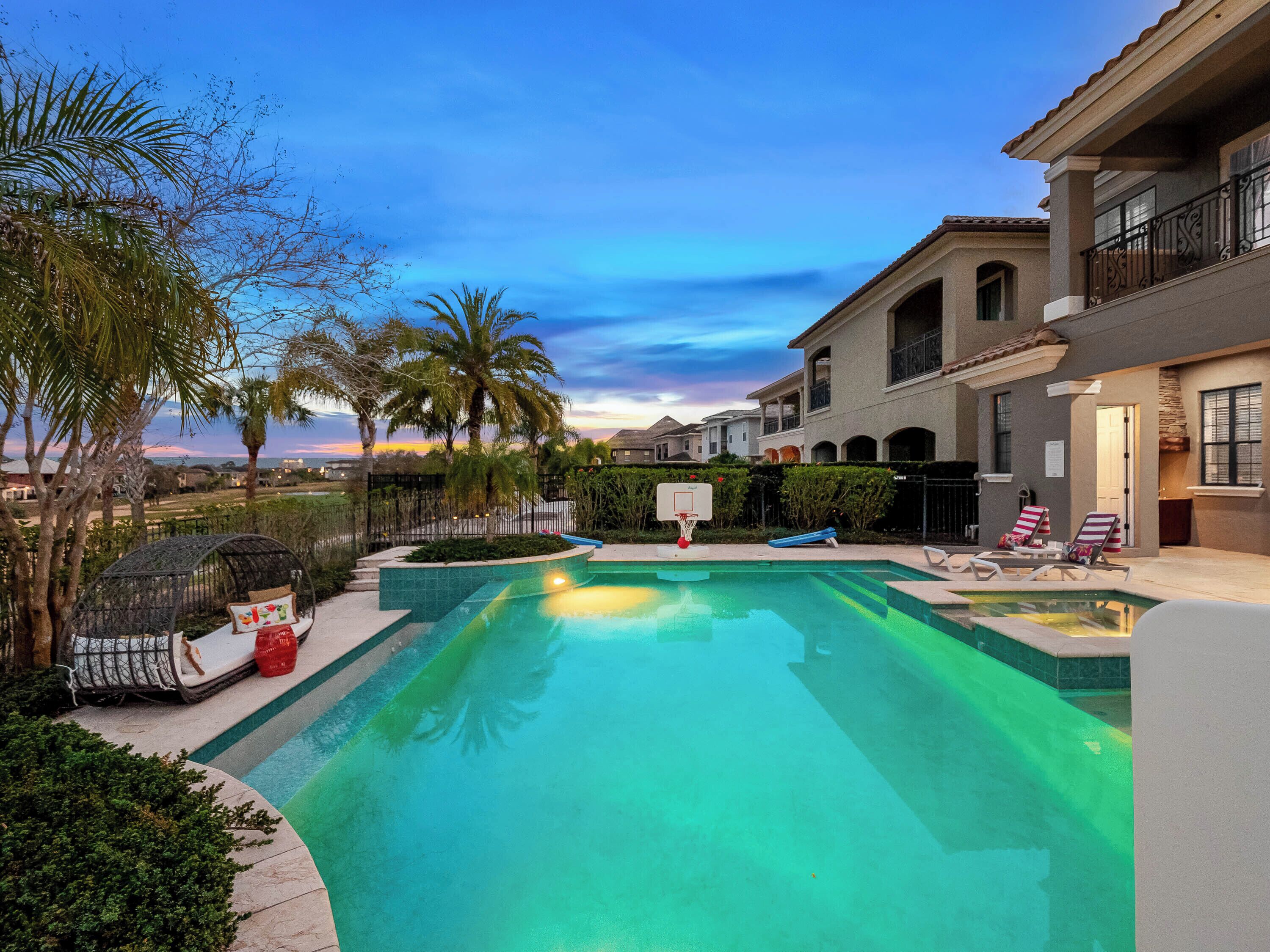 A swimming pool with a basketball hoop, palm trees, and a view of a residential area.