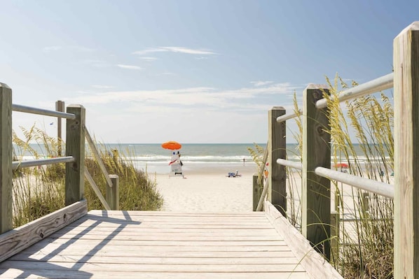 A wooden boardwalk leading to a beach with a person under an umbrella.