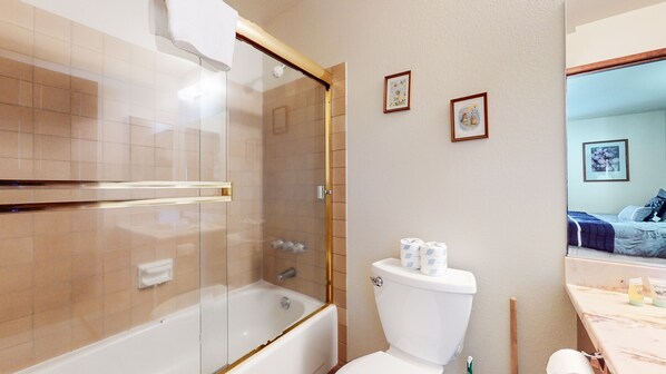 A bathroom with a bathtub, toilet, and a shower area. There are framed pictures on the wall and a bed visible through a mirror. - Spacious End Unit, Cozy Fireplace, Complex Hot Tub & Steps To Winter Shuttle (Mammoth Lakes)