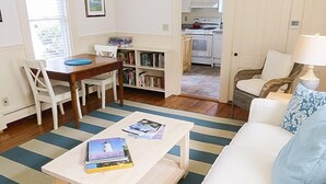 Ett mysigt vardagsrum med en randig matta, en vit soffa, ett träbord med böcker och en matplats med ett träbord och stolar. Köket syns i bakgrunden.