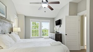Une chambre à coucher comprenant un lit, un ventilateur de plafond, un téléviseur, une fenêtre avec des stores et une porte.