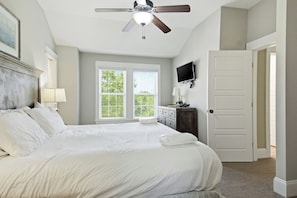 A bedroom with a bed, a ceiling fan, a TV, a window with blinds, and a door.
