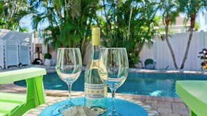Escena junto a la piscina con una botella y dos vasos sobre una mesa, rodeado de follaje y una pileta.
