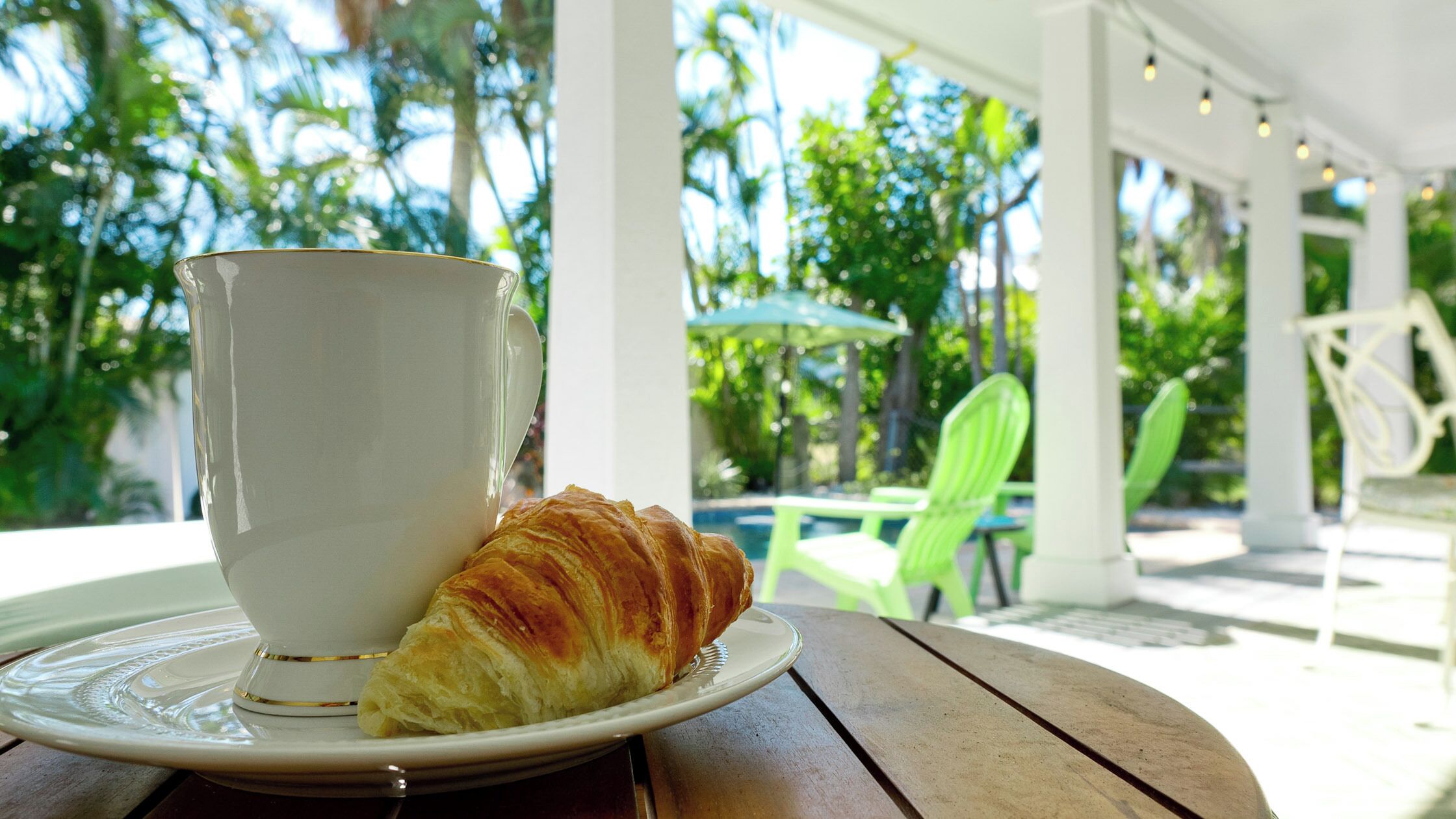 A cup of coffee and a croissant on a table in an outdoor setting with greenery and chairs.