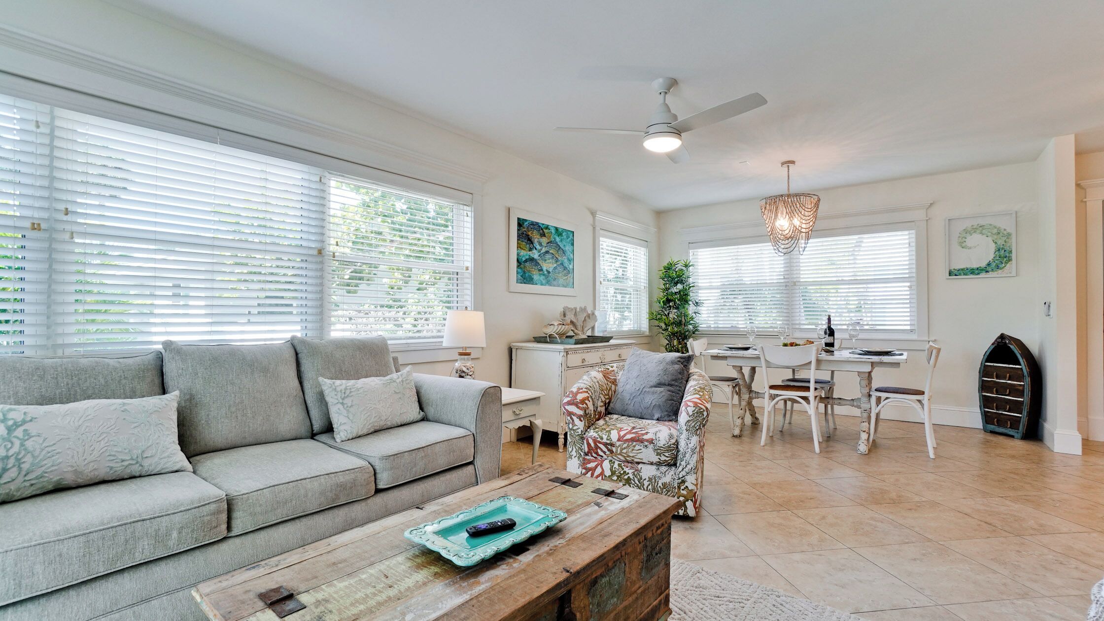 A living room with a grey sofa, a floral armchair, and a wooden coffee table. There is a dining area with a white table and chairs, and a ceiling fan above.