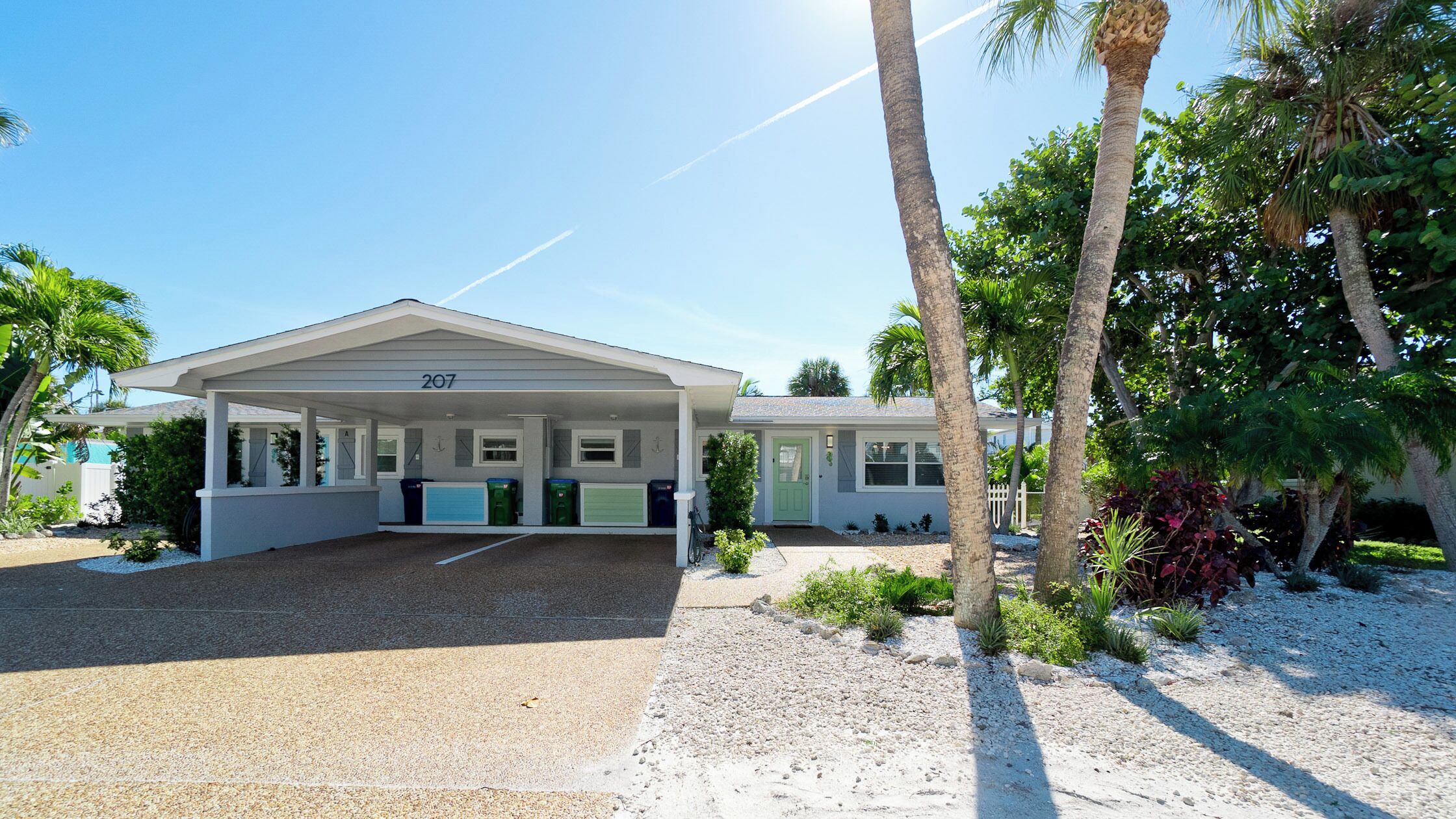 A single-story house with a covered porch, numbered 207, surrounded by palm trees and a gravel driveway.