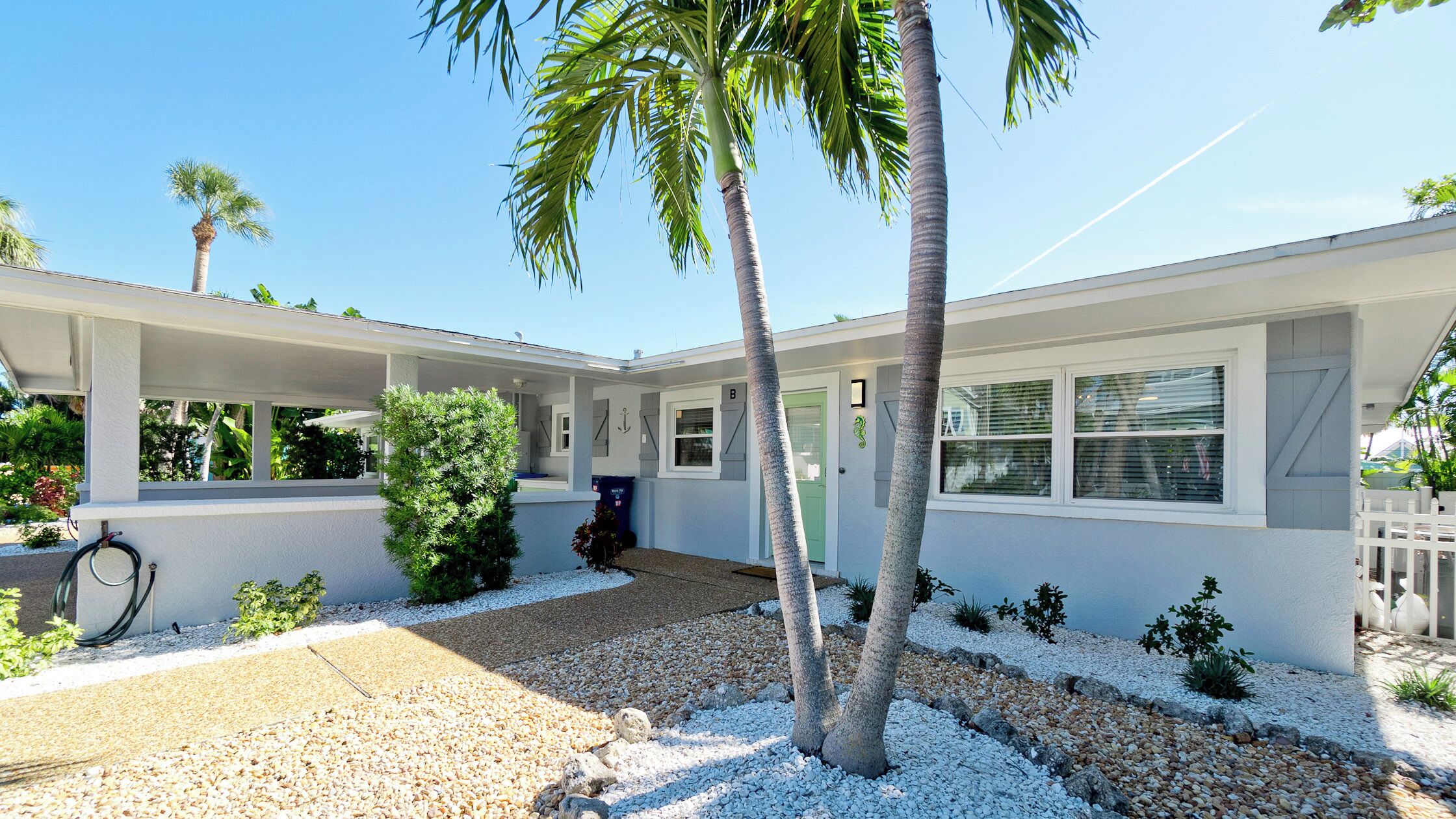 A modern two-story house with a covered porch, a small garden with palm trees, and a gravel driveway.