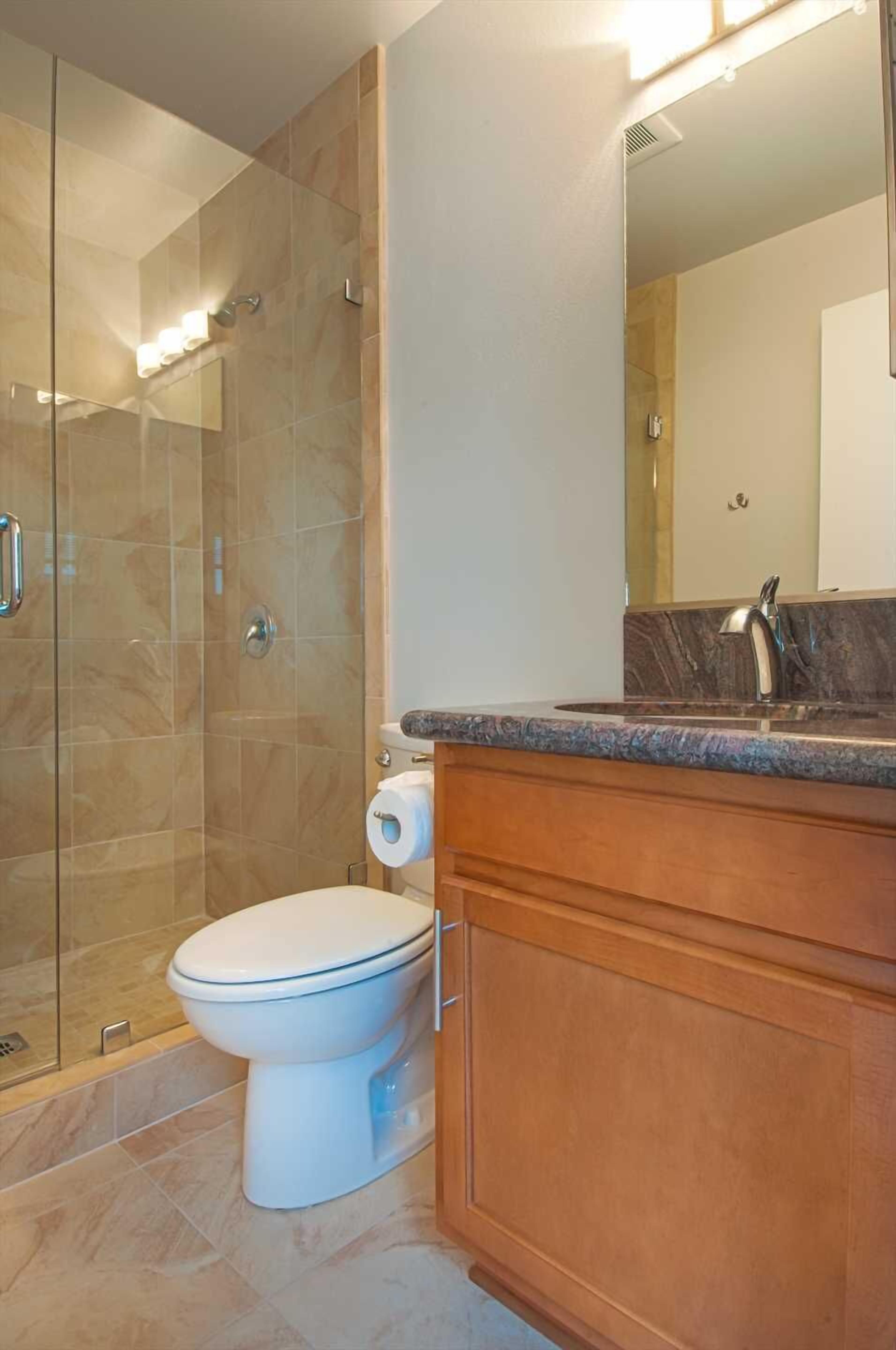 Un baño moderno con ducha de vidrio, inodoro blanco y tocador de madera con encimera de mármol.