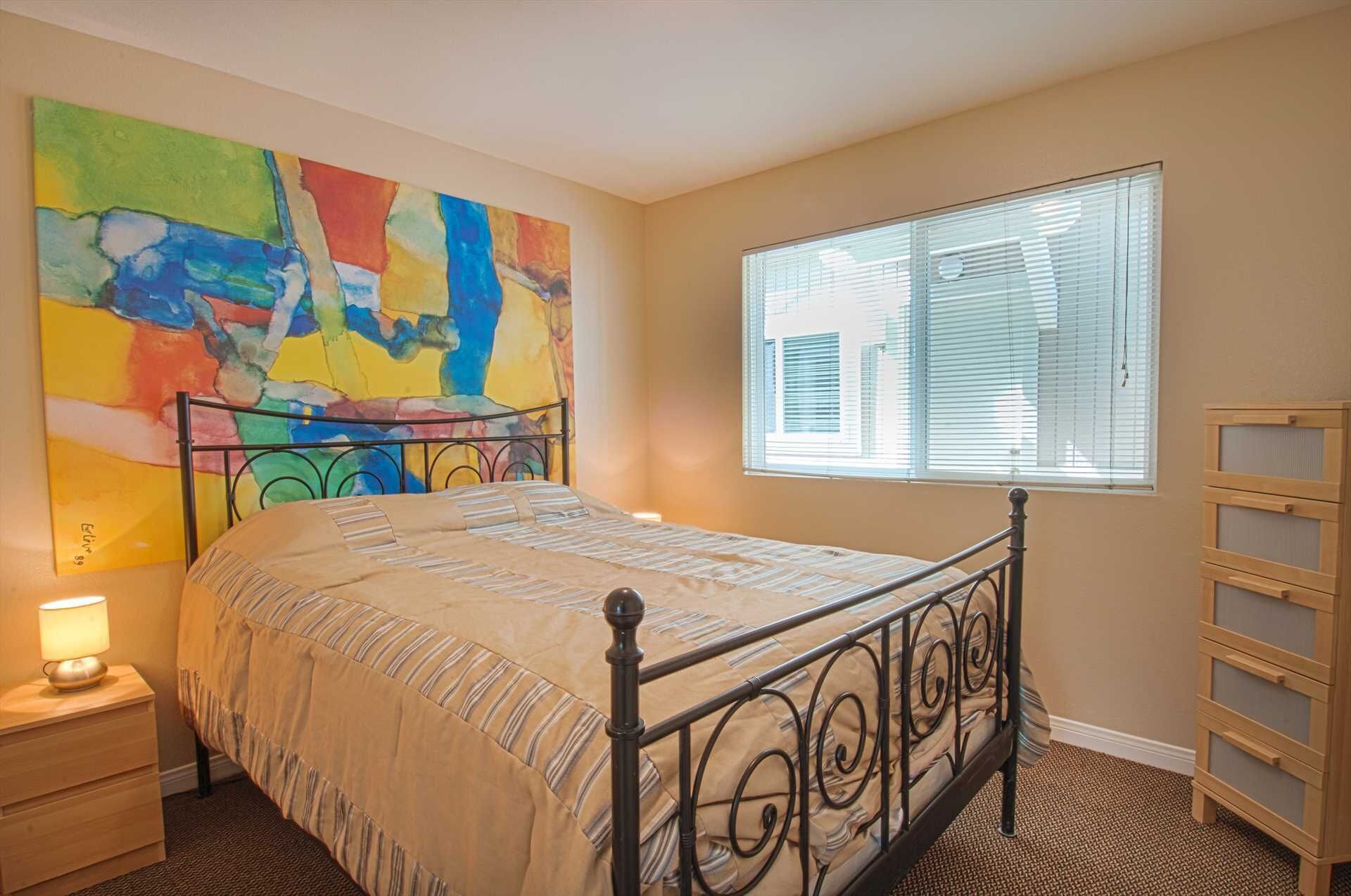 A bedroom with a bed, a nightstand, a window with blinds, a colorful abstract painting, and a wooden dresser.