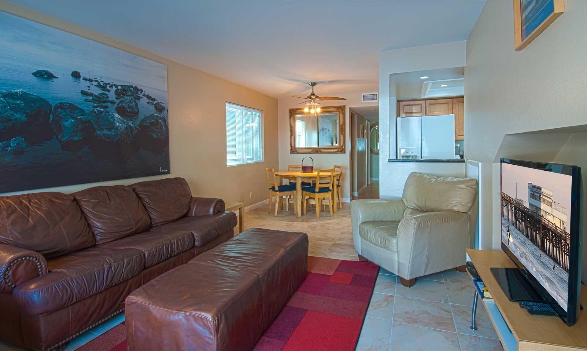 Una sala de estar con un sofá de cuero marrón, un sillón beige, una mesa de comedor con sillas y un televisor sobre un mueble.