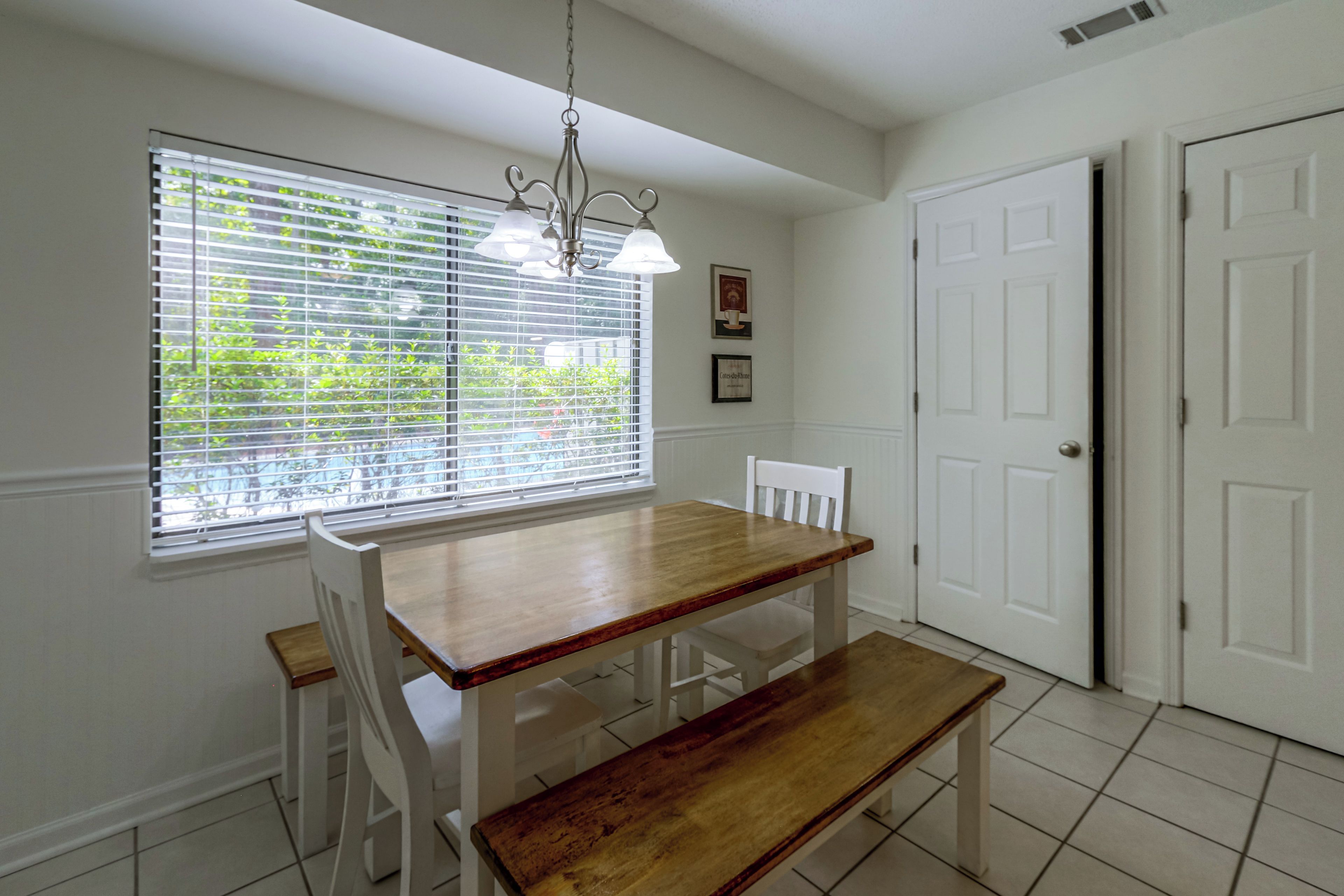 A dining area with a wooden table, white chairs, a bench, a hanging light fixture, and a window with blinds.