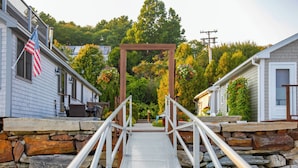 A metal bridge leading to a wooden archway in a residential area with stone walls and houses. - 'Heron Cottage' on Casco Bay w/ Deck & Boat Dock! (Freeport)