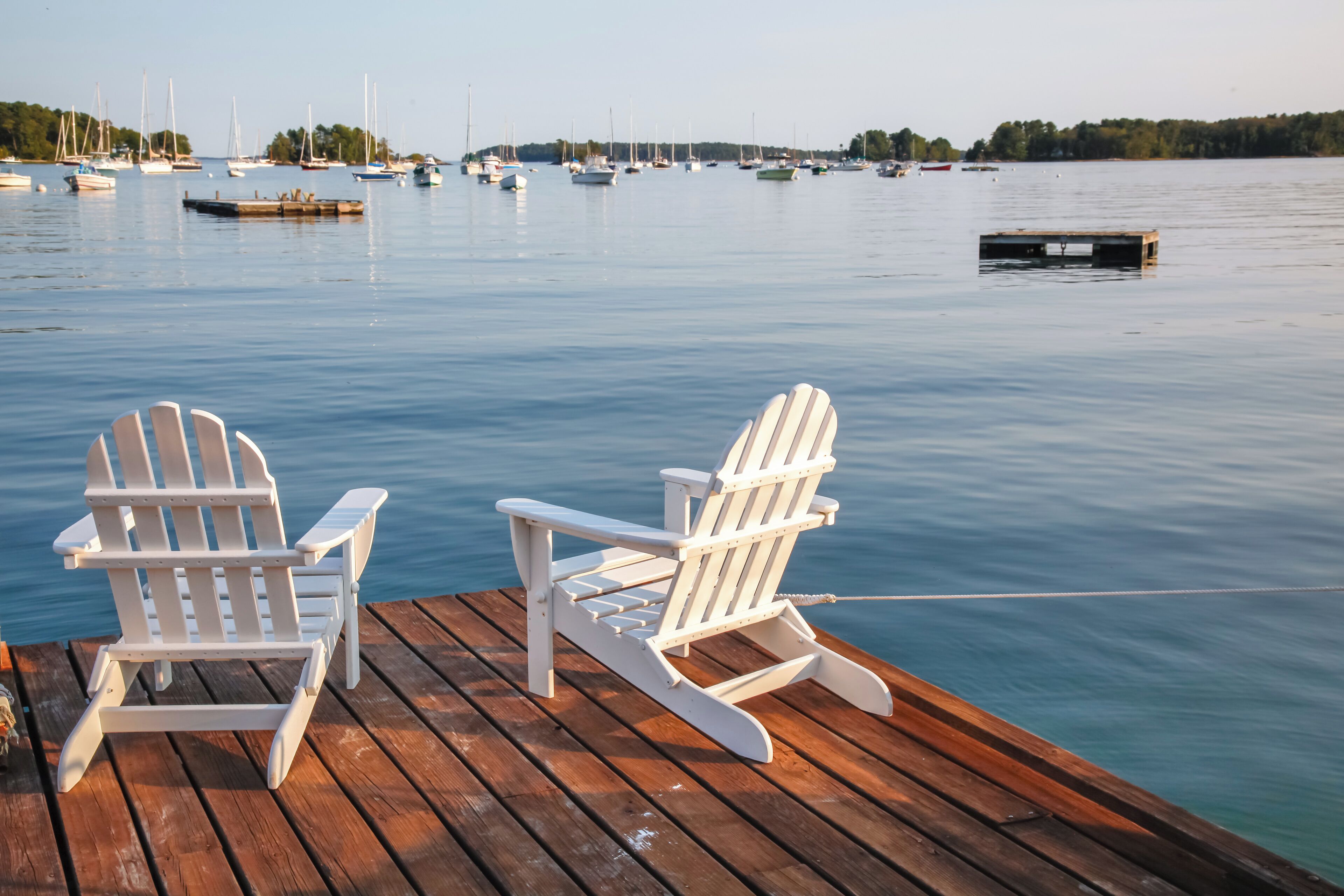Dos sillas Adirondack blancas sobre un muelle de madera con vistas a agua tranquila y varios barcos.