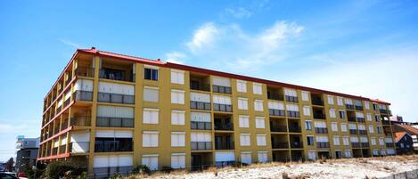 A multi-story hotel with a yellow facade and red roof, located on a sandy area with a wooden fence.