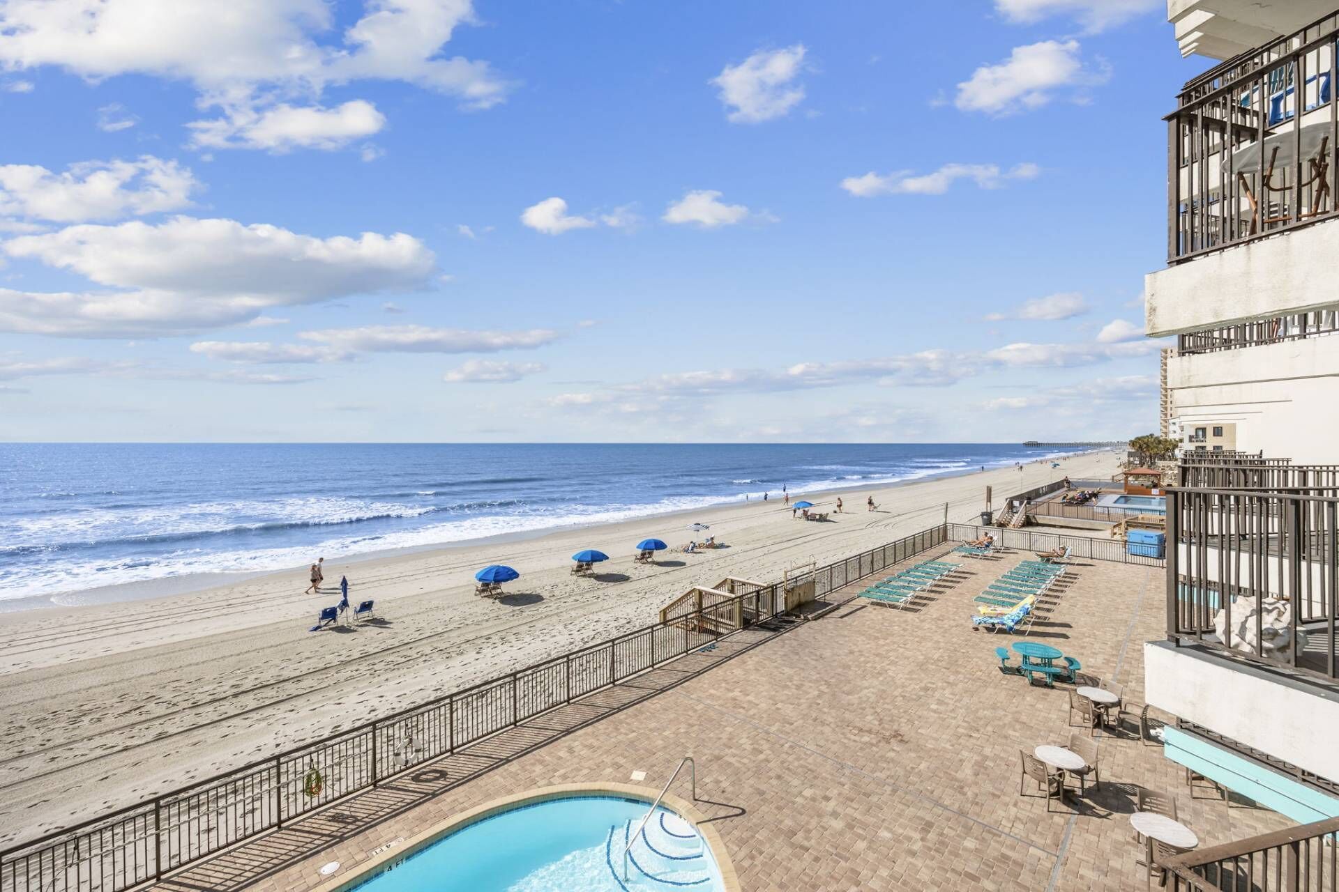 A beach view from a hotel balcony with a pool, lounge chairs, and umbrellas.