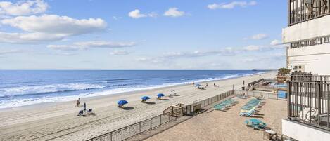 A beach view from a hotel balcony with a pool, lounge chairs, and umbrellas.