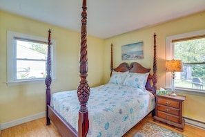 A four-poster bed with a wooden headboard, a bedside table with drawers, and a window with a view of the outside. - Lovely 'The Buoys Cottage' on Annisquam River! (Gloucester)