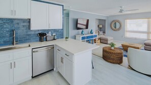 A modern kitchen with white cabinets, a dishwasher, and a blue backsplash. There is a dining area with a sofa, a coffee table, and a TV.