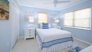 A bedroom with a bed, two bedside tables, two wall sconces, a ceiling fan, and a large window with blinds.