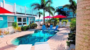 A swimming pool with blue chairs and umbrellas, surrounded by palm trees and a building with a blue facade.
