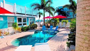 A swimming pool with blue chairs and umbrellas, surrounded by palm trees and a building with a blue facade.