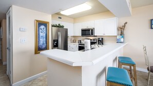 A modern kitchen with white cabinets, a stainless steel refrigerator, a microwave, and a coffee maker. There are two blue barstools and a vase with dried plants on the counter. - PI 412: Island Oasis - Enjoy a splashing good time at this beachfront condo! (Fort Walton Beach)