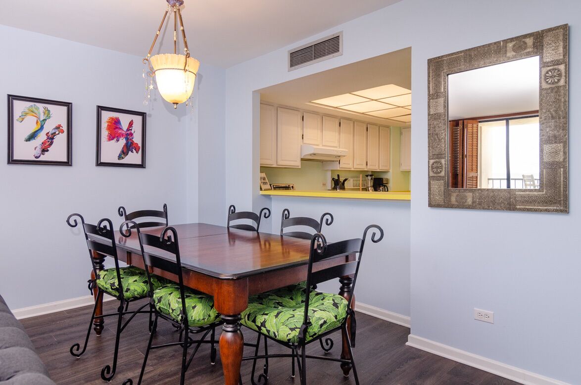 A dining area with a wooden table and chairs, a chandelier, framed artwork, and a view into the kitchen.